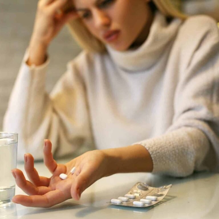 Frau mit Schmerzmittel in der Hand hat Rückenschmerzen und hält sich schmerzerfüllt den Kopf mit der rechten Hand