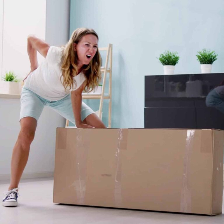 Chronische Rückenschmerzen - Akute Rückenschmerzen | Junge Frau die ein schweres Paket anheben will und dabei akute Rückenschmerzen kriegt