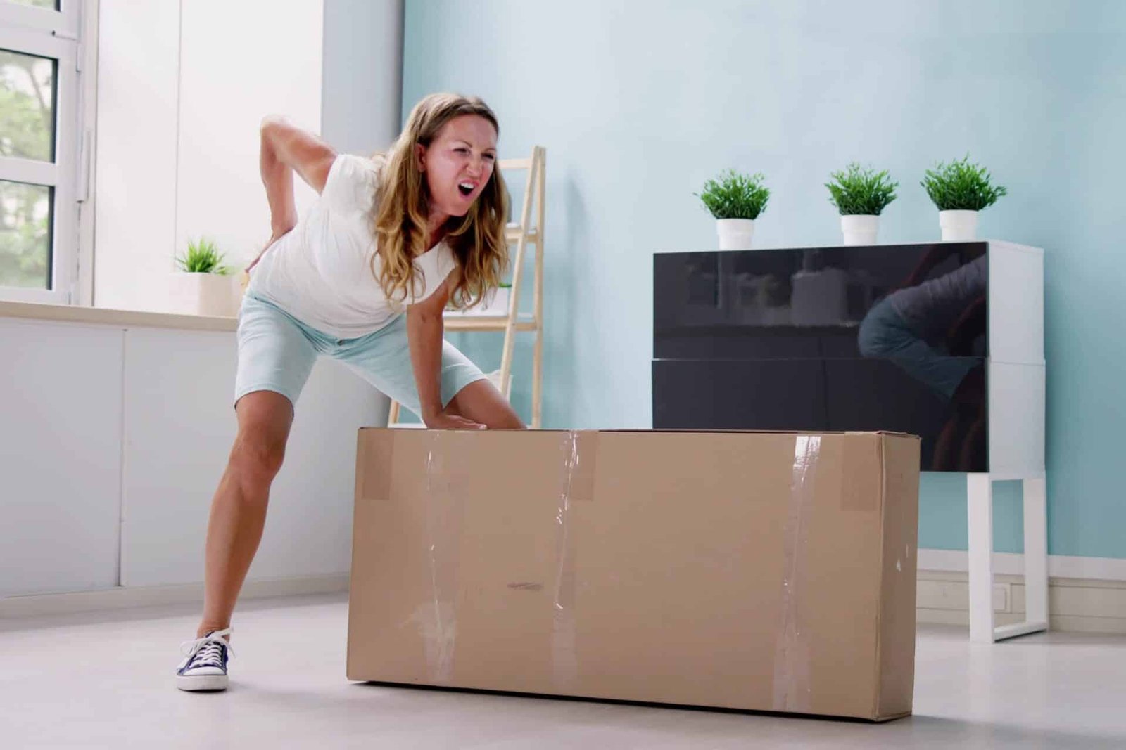 Junge Frau versucht ein schweres Paket anzuheben und bekommt akute Rückenschmerzen